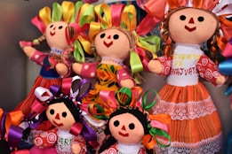 A collection of colorful handmade dolls with smiling faces and vibrant outfits. The dolls display various braided hairstyles with bright ribbon decorations. Each doll is uniquely dressed in traditional, intricately patterned clothing that adds charm and character to their appearance.