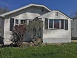 A small, single-story house with white siding and dark shutters. The front features a large window and a simple porch. There's a grass lawn with a few bushes, and a clear blue sky in the background.