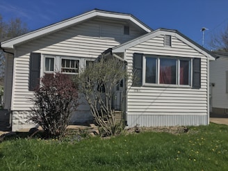 A small, single-story house with white siding and dark shutters. The front features a large window and a simple porch. There's a grass lawn with a few bushes, and a clear blue sky in the background.