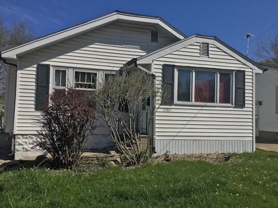 A small, single-story house with white siding and dark shutters. The front features a large window and a simple porch. There's a grass lawn with a few bushes, and a clear blue sky in the background.