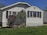 A small, single-story house with white siding and dark shutters. The front features a large window and a simple porch. There's a grass lawn with a few bushes, and a clear blue sky in the background.