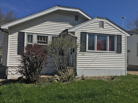 A small, single-story house with white siding and dark shutters. The front features a large window and a simple porch. There's a grass lawn with a few bushes, and a clear blue sky in the background.