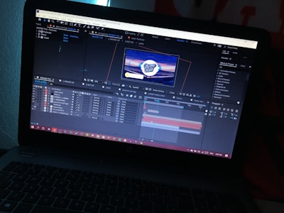 A laptop displays an open software program, likely a video editing or motion graphics application, showing multiple layers and a timeline with a preview of a logo in the center of the screen. The environment is dimly lit, highlighting the screen's glow.