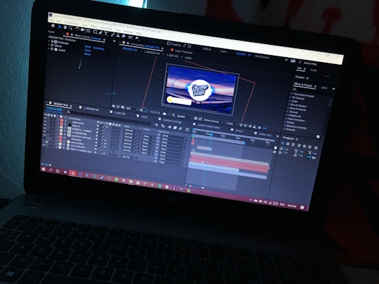 A laptop displays an open software program, likely a video editing or motion graphics application, showing multiple layers and a timeline with a preview of a logo in the center of the screen. The environment is dimly lit, highlighting the screen's glow.
