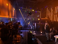 A music studio or concert setting with dim lighting and neon accents. Musicians are playing instruments like keyboards and guitars on a small stage. People stand and sit around the room, some with headphones or engaging in conversation, while cameras and recording equipment are visible.