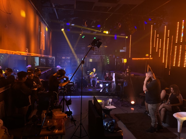 A music studio or concert setting with dim lighting and neon accents. Musicians are playing instruments like keyboards and guitars on a small stage. People stand and sit around the room, some with headphones or engaging in conversation, while cameras and recording equipment are visible.
