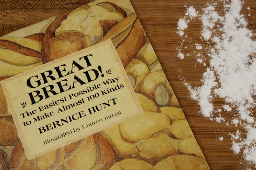 A cookbook titled 'Great Bread!' lies on a wooden countertop beside scattered flour. The book cover features images of various types of bread, hinting at its content about making different kinds of bread. The scattered flour adds a baking context to the scene.