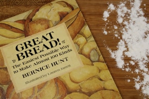 A cookbook titled 'Great Bread!' lies on a wooden countertop beside scattered flour. The book cover features images of various types of bread, hinting at its content about making different kinds of bread. The scattered flour adds a baking context to the scene.