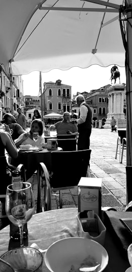 A bustling outdoor café scene with several people engaged in conversation. Tables are set with glasses and dishes. In the background, a historic square featuring a prominent equestrian statue and classic European architecture is visible. The setting has a casual, lively atmosphere under a large canopy.