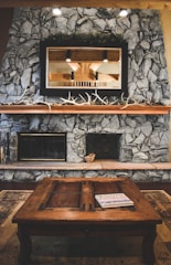 A rustic living room features a stone fireplace with a large wooden mantle, decorated with long antlers. Above the mantle is a mirror reflecting the interior beams of the room. In front of the fireplace, a wooden coffee table has a magazine or book placed on top, and a small woven basket sits on the hearth.