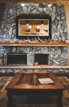 A rustic living room features a stone fireplace with a large wooden mantle, decorated with long antlers. Above the mantle is a mirror reflecting the interior beams of the room. In front of the fireplace, a wooden coffee table has a magazine or book placed on top, and a small woven basket sits on the hearth.