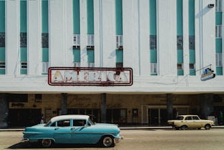 A vintage scene with two classic cars parked in front of a retro-style building. The building has a large sign reading 'America' in bold letters and a façade featuring vertical green stripes with small windows. The setting suggests a mid-20th-century or nostalgic atmosphere.