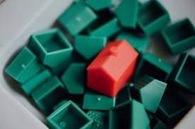 A collection of small, plastic green house pieces are scattered around and one slightly larger red plastic house is placed among them.