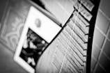 A close-up of a stringed instrument, likely a traditional Asian lute, placed against a tiled wall. The image is in black and white, highlighting the texture of the strings and the intricate details of the instrument's body. In the background, blurred shapes suggest books or framed art.