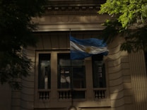 An elegant stone building with classical architectural elements, featuring a tall arched window and a prominent flag displayed in front. The Argentine flag, recognizable by its blue and white stripes with a sun emblem, is prominently waving. Surrounding greenery adds a touch of nature to the scene.