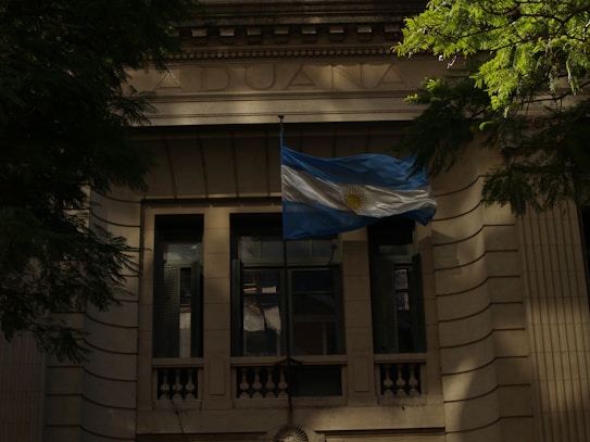 An elegant stone building with classical architectural elements, featuring a tall arched window and a prominent flag displayed in front. The Argentine flag, recognizable by its blue and white stripes with a sun emblem, is prominently waving. Surrounding greenery adds a touch of nature to the scene.