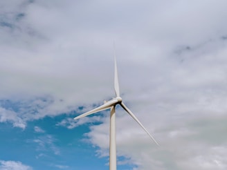 A tall wind turbine with three blades set against a partly cloudy sky. The turbine is prominently positioned, with its blades extending outwards against the backdrop of blue and white clouds.