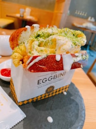 A richly textured egg sandwich placed in a decorative cardboard holder with 'The Eggbini' branding. The sandwich features fluffy scrambled eggs topped with a green herb garnish, nestled in a toasted bun. White sauce drips down the sides, adding to the dish's appeal. The background shows a casual dining setting with warm, wooden tones.