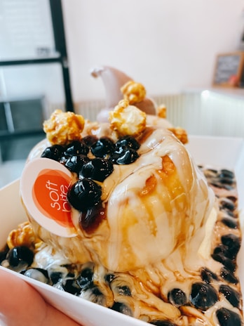 A dessert featuring a mound of ice cream drizzled with caramel sauce and topped with black tapioca pearls and caramel popcorn is presented in a white container. The texture of the ice cream appears creamy, and the overall presentation is indulgent, inviting, and artistic, capturing a sense of sweet indulgence.