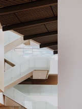 A modern interior architectural design featuring a series of staggered glass railings and stairs with wooden ceiling panels. The space is bright, with light streaming in from large windows in the background.