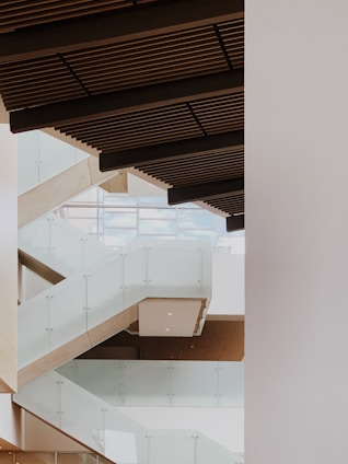A modern interior architectural design featuring a series of staggered glass railings and stairs with wooden ceiling panels. The space is bright, with light streaming in from large windows in the background.