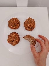 A hand is holding one of four oatmeal cookies on a white plate, with a bite taken out of it. The cookies have a rustic, homemade appearance with visible oats and a slightly chunky texture.