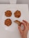 A hand is holding one of four oatmeal cookies on a white plate, with a bite taken out of it. The cookies have a rustic, homemade appearance with visible oats and a slightly chunky texture.