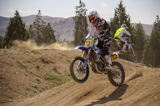 Two motocross riders are racing on a dirt track surrounded by tall trees and mountainous terrain. The riders, dressed in protective gear, are in action, with one in the foreground executing a jump, kicking up a cloud of dust. The scene conveys a sense of speed and adrenaline amidst a natural backdrop.