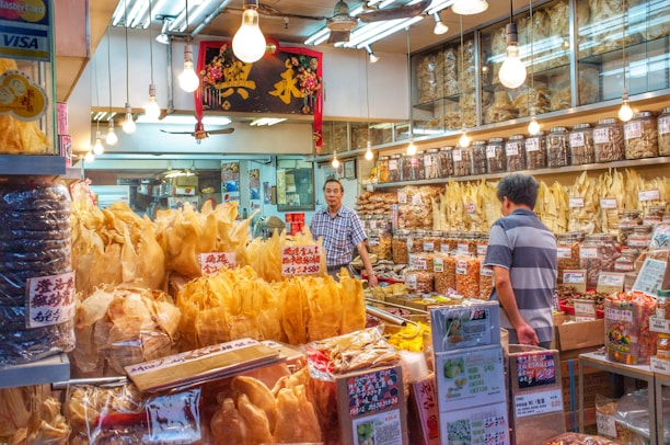 A traditional shop selling various dried goods, with shelves full of jars and containers. Two men interact in the brightly lit store, surrounded by an array of packaged products and handwritten signs displaying prices. Hanging light bulbs add a warm glow to the bustling environment.