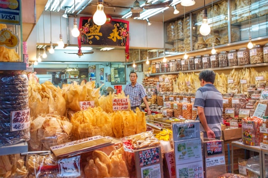 A traditional shop selling various dried goods, with shelves full of jars and containers. Two men interact in the brightly lit store, surrounded by an array of packaged products and handwritten signs displaying prices. Hanging light bulbs add a warm glow to the bustling environment.
