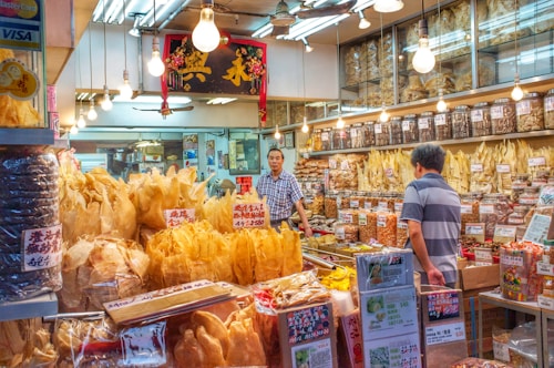 A traditional shop selling various dried goods, with shelves full of jars and containers. Two men interact in the brightly lit store, surrounded by an array of packaged products and handwritten signs displaying prices. Hanging light bulbs add a warm glow to the bustling environment.