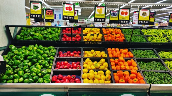 A colorful arrangement of various types of capsicums displayed in a grocery store, including green, red, yellow, and orange varieties. Each variety is neatly organized in separate trays with price tags above them. The background features signs and advertisements that indicate the location in a produce section.