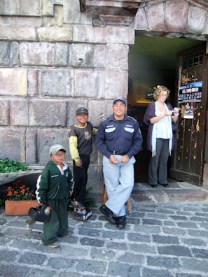 A group of people, including two children, a security guard, and a woman, are standing in front of a stone building entrance. One child is holding shoe shine supplies. There are potted flowers on the ground.