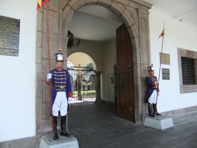 Two uniformed guards stand at attention outside an arched doorway. They wear blue jackets with red accents, white pants, tall black boots, and shiny helmets. Each holds a tall ceremonial staff adorned with flags. The architecture includes stone elements and decorative plaques on the walls.