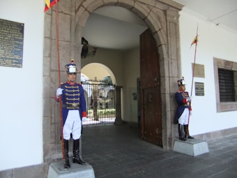 Two uniformed guards stand at attention outside an arched doorway. They wear blue jackets with red accents, white pants, tall black boots, and shiny helmets. Each holds a tall ceremonial staff adorned with flags. The architecture includes stone elements and decorative plaques on the walls.