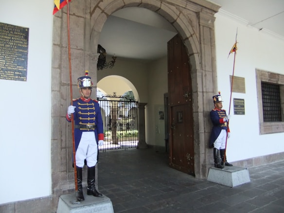 Two uniformed guards stand at attention outside an arched doorway. They wear blue jackets with red accents, white pants, tall black boots, and shiny helmets. Each holds a tall ceremonial staff adorned with flags. The architecture includes stone elements and decorative plaques on the walls.