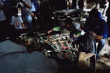 A group of people gather around a table displaying various small, colorful figurines. The figures resemble animals and other creatures, and are arranged closely together. The scene is partially shaded, with sunlight filtering through nearby trees. Some individuals are intently observing, while others seem engaged in handling or discussing the items.