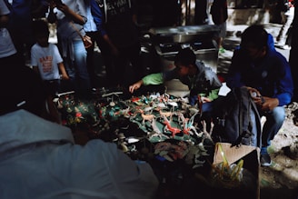 A group of people gather around a table displaying various small, colorful figurines. The figures resemble animals and other creatures, and are arranged closely together. The scene is partially shaded, with sunlight filtering through nearby trees. Some individuals are intently observing, while others seem engaged in handling or discussing the items.