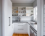 A modern kitchen features white cabinetry and shelves filled with neatly arranged dishes and glasses. The backsplash consists of white subway tiles, and a stainless steel mixer is placed on the countertop. A stainless steel sink and dishwasher are also visible.