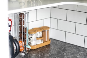 A kitchen countertop features an organized setup with a vertical metal rack holding coffee pods, and a wooden tray containing jars with cork lids for storing tea bags, sugar, and other kitchen essentials. There is also an electric kettle partially visible on the left side. The backsplash is tiled with white rectangular tiles and the countertop has a dark marbled pattern.