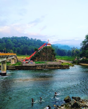 A serene riverside landscape featuring a small temple perched on a lush hill, with a long red staircase leading up to it. The river flows gently in the foreground, where three people are wading in the water. A bridge crosses over part of the river, and dense greenery fills the background, with misty mountains visible in the distance.
