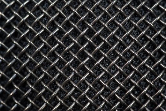 A close-up view of a metallic mesh pattern with interwoven wires forming a grid-like structure. The background appears dark, highlighting the reflective quality of the silver-colored metal.