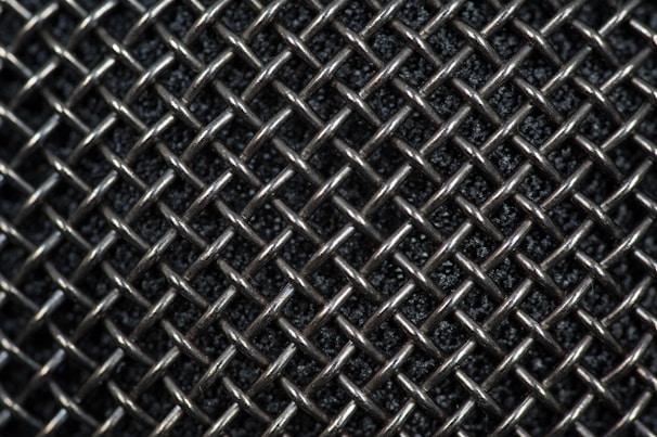 A close-up view of a metallic mesh pattern with interwoven wires forming a grid-like structure. The background appears dark, highlighting the reflective quality of the silver-colored metal.