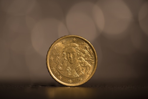 A detailed view of a gold-colored coin standing upright against a blurred, warm-toned background. The coin features an intricate design of a face with flowing hair surrounded by stars and the year 2002 with the letters 'R' on it.