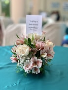 A floral arrangement featuring white and pink flowers with green foliage, placed on a teal tablecloth. Above the arrangement is a card with a motivational Spanish quote.