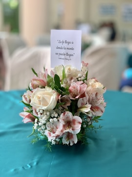 A floral arrangement featuring white and pink flowers with green foliage, placed on a teal tablecloth. Above the arrangement is a card with a motivational Spanish quote.