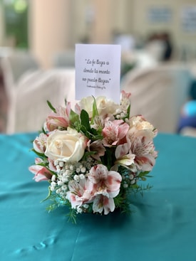 A floral arrangement featuring white and pink flowers with green foliage, placed on a teal tablecloth. Above the arrangement is a card with a motivational Spanish quote.