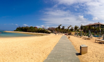 A picturesque beach setting featuring a sandy shoreline with a paved pathway leading to the distance. Lush green vegetation and a red-roofed building are visible on the left, while palm trees and lounge chairs line the right side, next to a modern building. The sky is bright blue with scattered clouds.