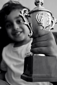A smiling child holding a silver trophy in one hand. The trophy is intricately designed with ornate handles and a shiny surface, indicating a sense of accomplishment or victory. The child is wearing a simple light-colored shirt.