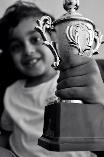 A smiling child holding a silver trophy in one hand. The trophy is intricately designed with ornate handles and a shiny surface, indicating a sense of accomplishment or victory. The child is wearing a simple light-colored shirt.